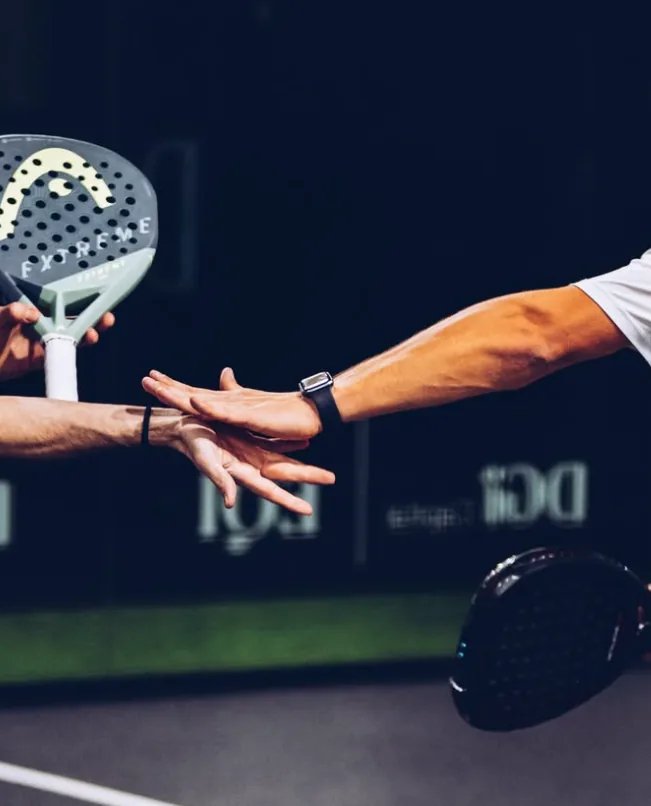 Dois jogadores se cumprimentam com as mãos em quadra, com raquetes de padel e a placa DGI / EQI ao fundo.